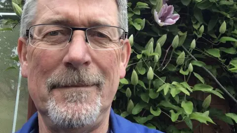 A man wearing glasses and a blue top, with short grey hair and a goatee beard, stands in front of a climber plant with a purple flower.