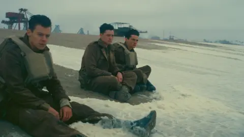 Warner Bros Harry Styles sits on the beach with water lapping his feet, with two other actors also dressed in military uniform to his left.
