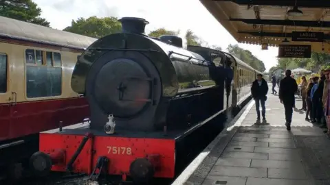 A black steam train waiting at a platform. There are people on the platform to the right of the train. To the left is a train carriage. The sky is cloudy. 