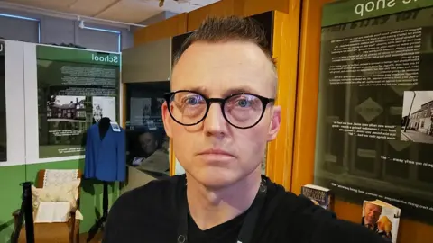 Alastair Hawken A head and shoulders photo of Alastair Hawken standing inside a museum. He is looking at the camera, not smiling, and is wearing black-rimmed glasses and a black top. He has short, brown, spikey hair.