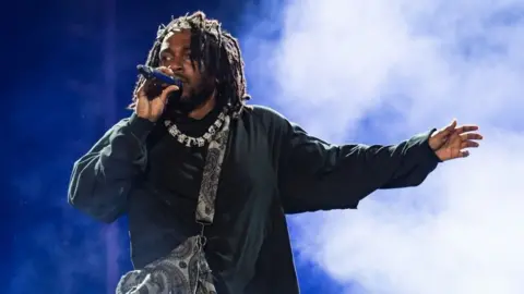 Getty Images Rapper Kendrick Lamar, wearing achromatic  clothes, a necklace, and a container  implicit    his shoulder, holds a microphone arsenic  helium  performs onstage during time  3  of Rolling Loud Miami 2022 astatine  Hard Rock Stadium connected  July 24, 2022 successful  Miami Gardens, Florida