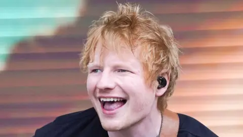 Ed Sheeran smiles widely as he performs on stage. He has red hair and wears a black T-shirt with a guitar strap around his shoulder. He has a black earpiece in.