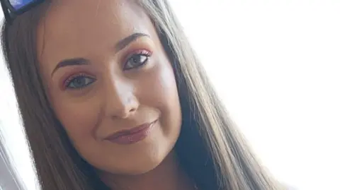 A close-up photo of Chloe Mitchell - a young woman with long, straight, fair hair - smiling at the camera. She is wearing black-framed sunglasses with blue lenses on her head, which are partially visible. 