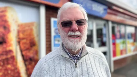 An elderly man with short white hair, a white beard and dark glasses is wearing a check shirt under a grey sweater.