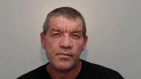 GMP Mugshot of Keith Bretherton,. He has short grey hair, a five o'clock shadow and is wearing a black t-shirt.