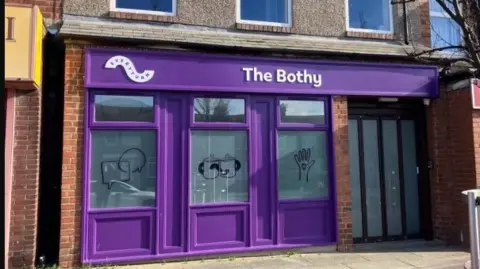 Everyturn Mental Health The Bothy is a purple fronted building with a door to one side and three windows, one with various drawings like a hand in the air, and two cups of tea 