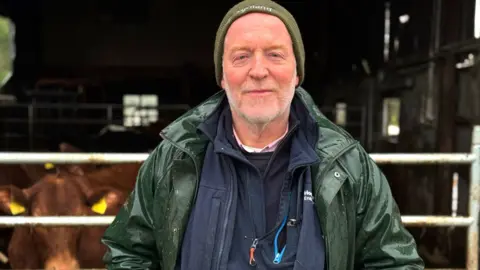 Richard Halleron is an older man. He is standing in front of a shed with metal gate which is closed in front of some brown cows. He has a short white beard and is wearing a green beanie hat, along with a dark green rain coat over a navy blue zip up top. 