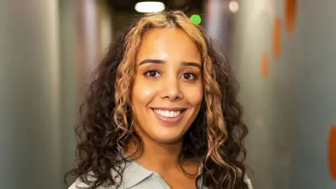 Ending Violence Against Women Janaya Walker smiles at the camera. She has curly, medium length brown hair with some lighter blonde pieces that frame her face. 