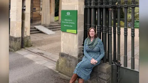 Fiddler sitting by the Arnos Vale Cemetery gates. She is wearing a blue and white triangular patterned dress with a green-grey shawl and brown boots. She has shoulder length brown hair and is smiling at the camera with her hands held together on her lap. The black metal gates are directly behind her and there is a green sign sayign 'welcome to Arnos Vale Cemetery'.
Behind the gates is a walkway and a ramp up onto a side bit. In the background there are trees and grass.