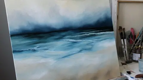 Anna Corbishley A stormy picture of the sea and shore painted on a square canvas, propped up on an easel. There are dark blues on the horizon, and browns on the bottom for the sand. There is a pot of paintbrushes on the table on the right