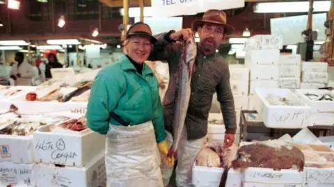 Two traders stand in front of crates of different types of fish