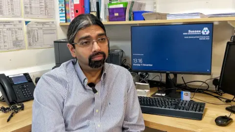 BBC/Mark Norman Dr Bikram Raychaudhuri sits at his desk in a GP surgery