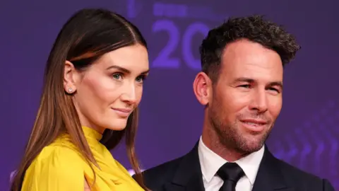 Mark Cavendish has curly dark hair and is wearing a black suit jacket, white shirt and black tie. Peta Cavendish has long dark hair and is wearing a yellow dress. Both are posing at an awards evening.