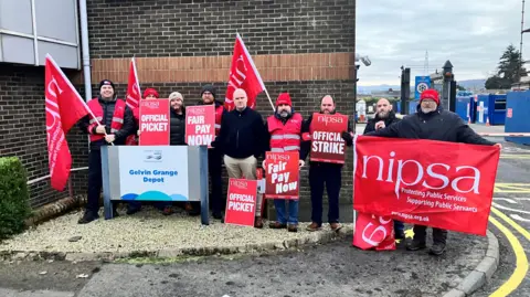Nine men stand in front of a black and red brick building. All the men are holding either a red flag or red placard. The signs and flags say 'Nipsa,' on them in red font. Some signs say 'Official picket' others say 'Fair pay now'.