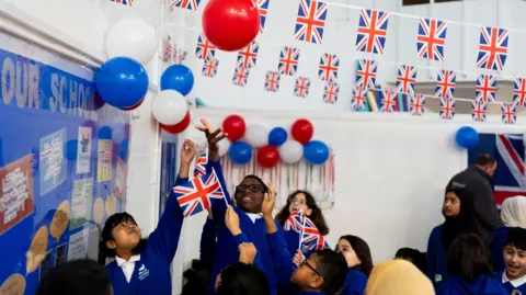 PA Media School children dressed in blue reaching up to touch balloons, some holding union flags 