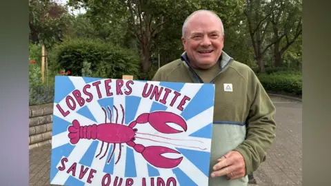 A middle-aged man in a khaki fleece is smiling to camera and holding a placard which says 'Lobsters unite, save our lido' with a red painted lobster on a blue and white striped background. The man is on a driveway with trees behind