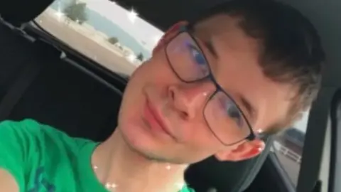 Daniel Peake A close-up of Daniel sitting in a car and smiling to camera. He is wearing black glasses and a green t-shirt