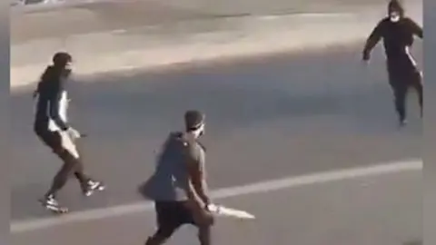 Three men wearing hats and/or masks confront each other in the street. They all appear to be holding knives/bladed weapons.