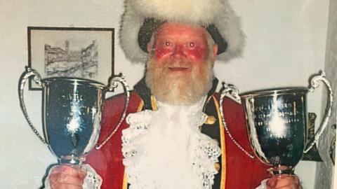 Mr Heeks pictured after winning the European Championships. He is holding two large silver trophies, and wears a bright red jacket and a large fur-lined hat. 