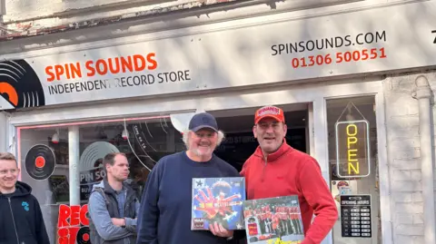 Ali Timson / BBC Two men holding record sleeves stood outside a record shop with people queing behind them.