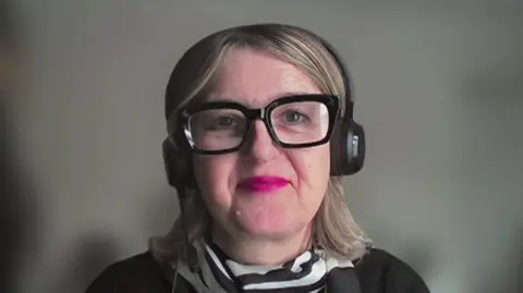 A woman with blonde shoulder length hair and chunky black glasses smiles to camera with a blurred background. She is wearing bright pink lipstick, a white and black blouse and black jacket.