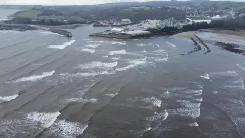 An aerial view of a bay with a harbour in the distance and a beach