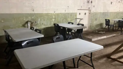 An underground space is seen with classroom tables and chairs in it. The walls are unfinished and appear to be made of concrete.