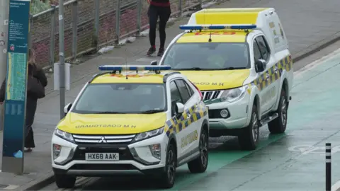 Supplied Two four-by-four vehicles parked by the side of a road. They have a yellow and blue livery and HM Coastguard is written across their bonnets.