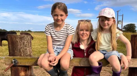 Heather Oldfield stands in the centre of the image wearing a burgundy fleece gilet and sunglasses on her head. On either side of her is a young child. They are in a field with cattle behind them.