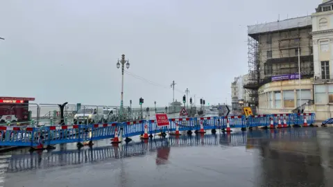 A section of blue barriers across the road which leads to the hotel which can be seen covered in scaffolding on the right of the image. It is a grey and wet day with the sea just visible in the background.