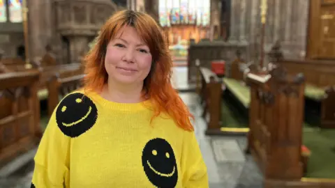 A woman with red hair in a yellow jumper with black smiley faces on it. She is standing in front of wooden pews and is smiling at the camera