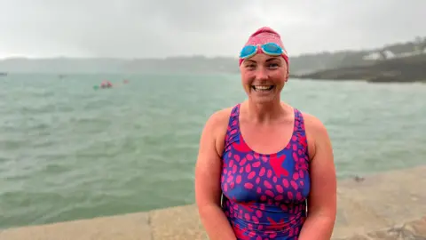 A person in a pink and purple patterned swimsuit and pink swim cap stands on a concrete surface near choppy water, under an overcast sky. Distant swimmers and a shoreline with greenery are visible in the background.