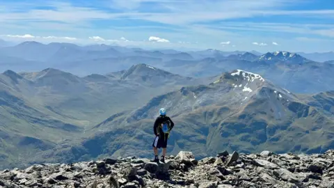 A man stands at the summit of Ben Nevis with a dramatic view of adjoining hills