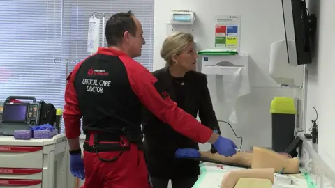 The Duchess of Edinburgh with a critical care doctor at the facility during a training session. The doctor is with his back to the camera. Sophie is looking up at a screen. Equipment could be seen everywhere in the room.