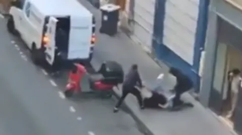 A still from a video shows two men in black trying to drag a woman into their white van