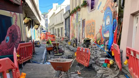 Guernsey Water A narrow street lined with colourful graffiti is seen with the cobbles lifted and placed to the side, orange barriers in place and wheelbarrows, a broom and other building materials scattered along the way. 