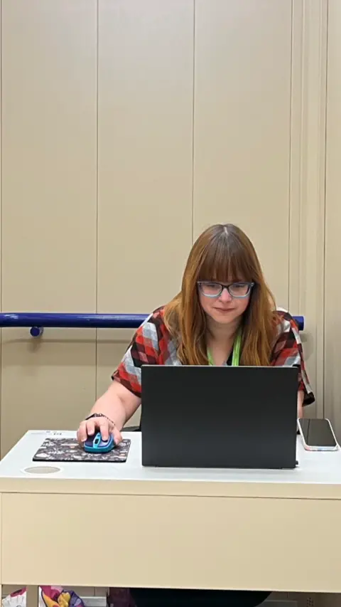 A woman with brown hair and a fringe sitting behind a laptop at a desk. She has set up her own personal office inside a lift.