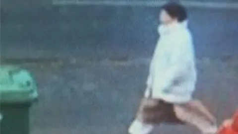 Missing People A woman in a light-coloured coat walking on a street in a blurry CCTV image.
