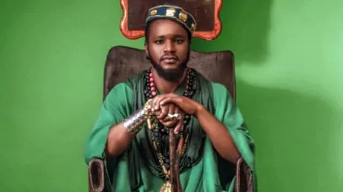 @aamronblack em tiktok aamron retratado sentado em uma cadeira de madeira lindamente esculpida, vestindo um vestido verde, chapéu verde decorado, muitas miçangas e outras jóias. Suas mãos descansam em uma bengala cerimonial. Atrás dele, o fundo é verde brilhante.