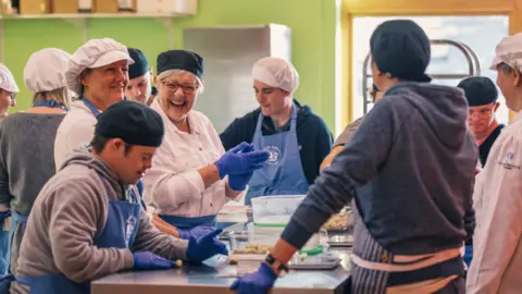 Step and Stone Students and staff baking together with smiles on their faces. They are wearing blue hygiene gloves, aprons and hair caps while shaping dough. They have a big commercial style kitchen top they are working on and there is a big fridge behind them. There are a few other people working at a different counter top to the left of the picture.