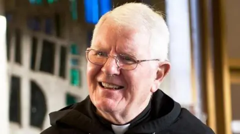 Bishop Michael Campbell in a black habit in front of a stained glass window