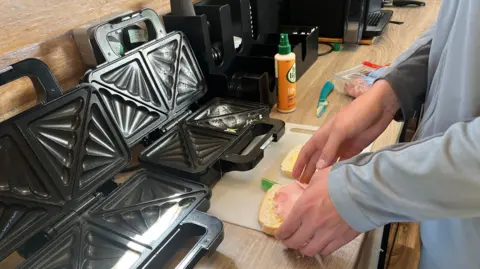 John Devine/BBC A sandwich is being prepared on a work surface. Ham is being put on cheese on a slice of bread. There are toastie machines on the side.