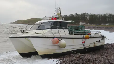 The Sea Eagle has a catamaran-like hull with a small cabin and there are green creels on the desk. The boat is on a shingle beach.
