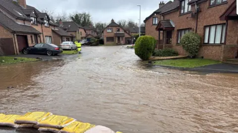 Flood water on a street in Culcavy, with yellow sand bags in front. There are houses and cars in the background.