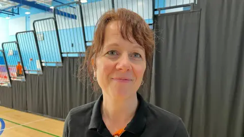 Ant Saddington/BBC A woman in a black polo stands by the viewing area off court smiling at the camera.