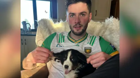 Family picture A young man with brown hair and stubble, wearing a Donegal jersey, holds a border collie puppy on his knee. The puppy is mid yawn and its tongue is out. He gently holds the puppy's ears with both hands, stretching them apart.