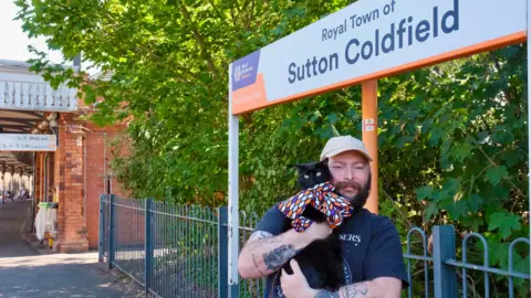 On a railway station platform is an orange, white and purple sign which says Royal Town of Sutton Coldfield. Underneath is a man wearing a beige cap. He has a black beard and tattoos. In his arms is a black cat with a multicoloured scarf on. 