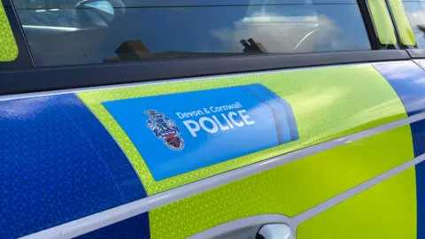 A generic image of the side of a police car which says 'Devon and Cornwall Police' on it. The car door is yellow and blue.