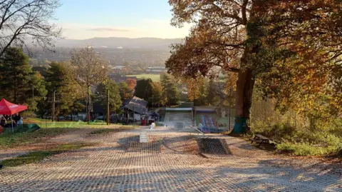 A view from the midway point of an artificial ski slope and ramp. There are trees to both sides and a landscape of hills, houses and a field in the background.