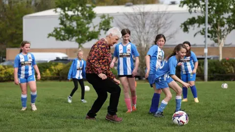 England Football A former Lioness is seen playing football on a pitch against a local team. She is wearing a black and red top, and black trousers. The girls are in a blue and white strip. 